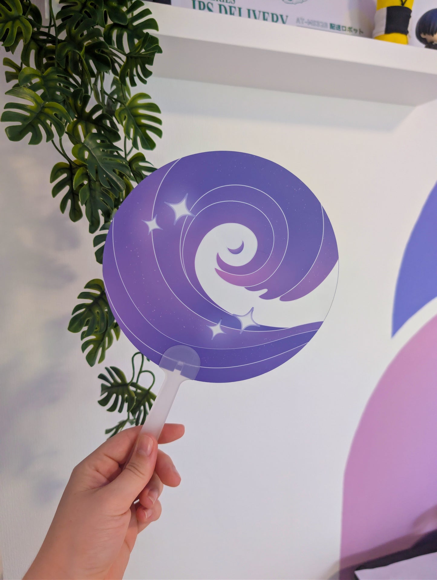 A hand holds a round clear plastic fan with a purple spiral lollipop design featuring white stars and a crescent moon, set against a white wall with a green plant on the left.