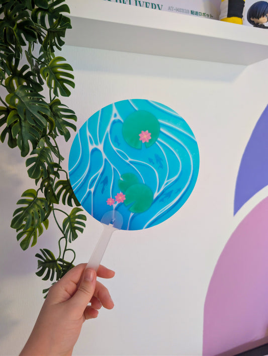 A hand holding a round paper fan with a blue and green water ripple pattern and pink lotus flowers, positioned next to a green plant in front of a white wall.